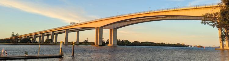 A low set concrete bridge spans a vast river. A man and child stand on the jetty.